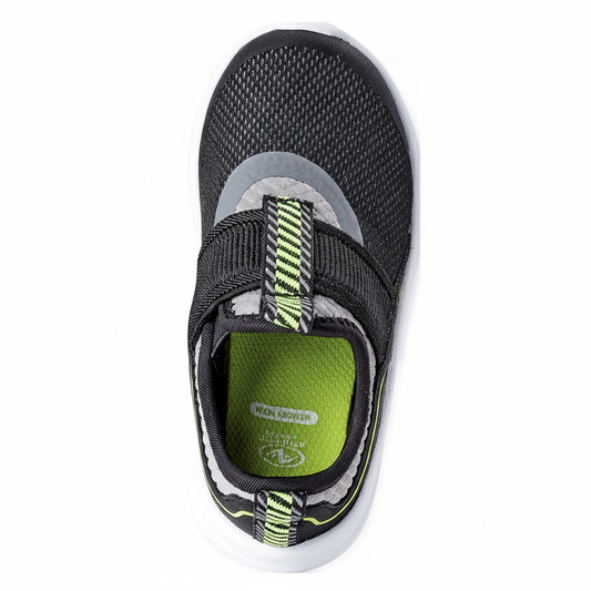 Angled 3/4 top view of kids black slip-on sneaker showing mesh upper, neon pull tab and green memory foam insole on clean white background